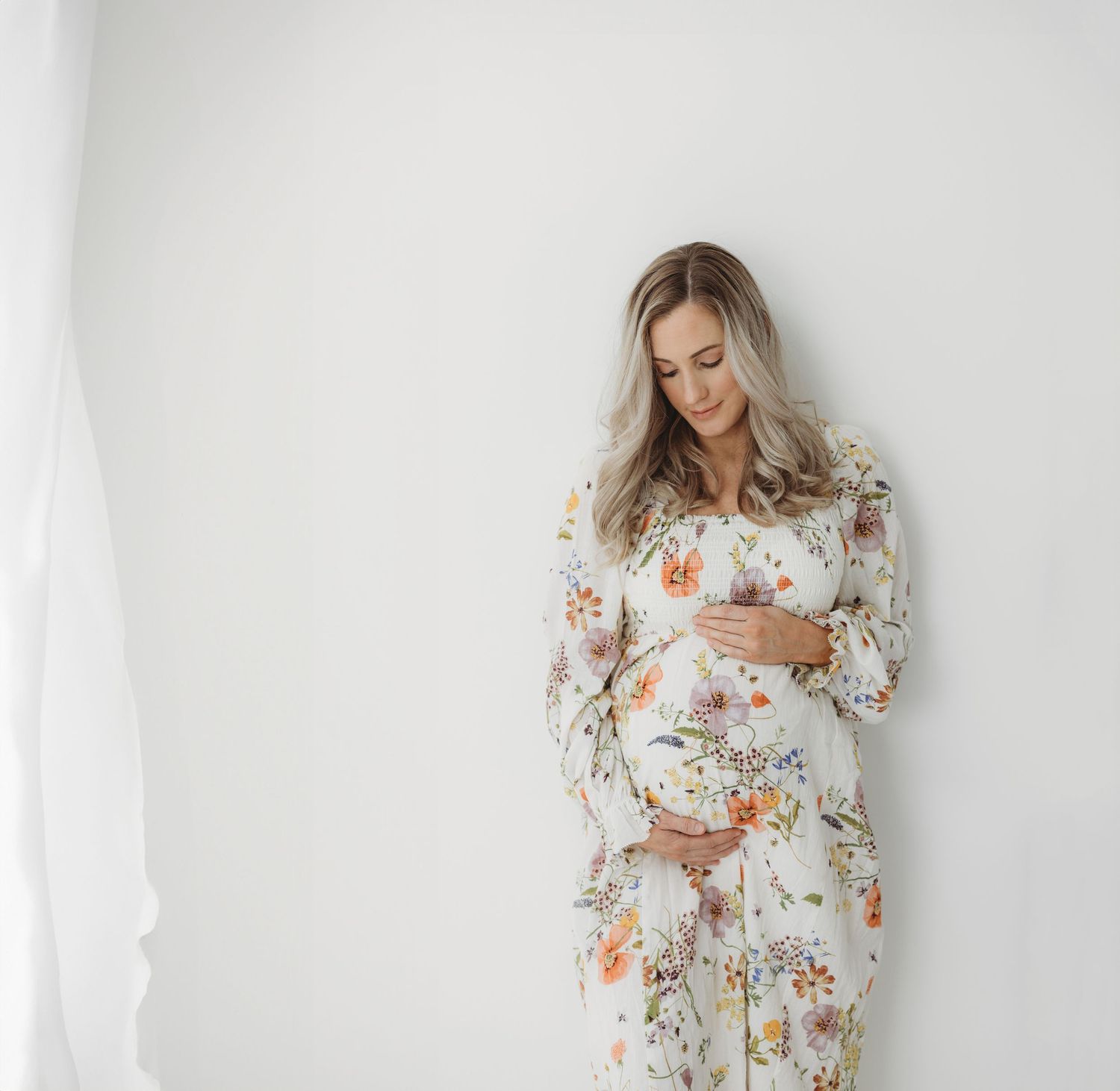 Expectant figure in floral dress touches belly against white wall.