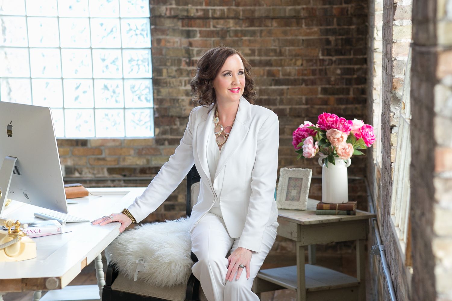 Stylish woman in white suit leans against desk in bright rustic brick loft office space.