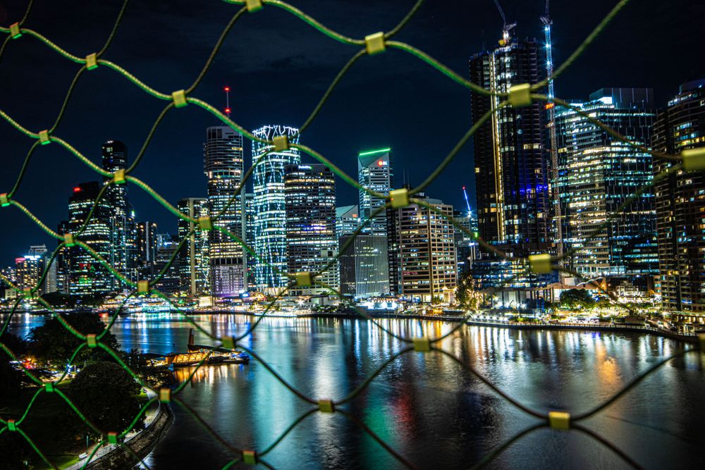 Brisbane's illuminated downtown skyline reflected in the river, viewed through safety mesh.