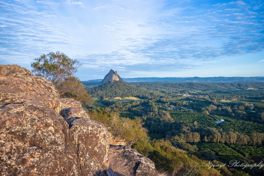View of Glass House Mountains from scenic outlook with blue sky and rugged terrain.