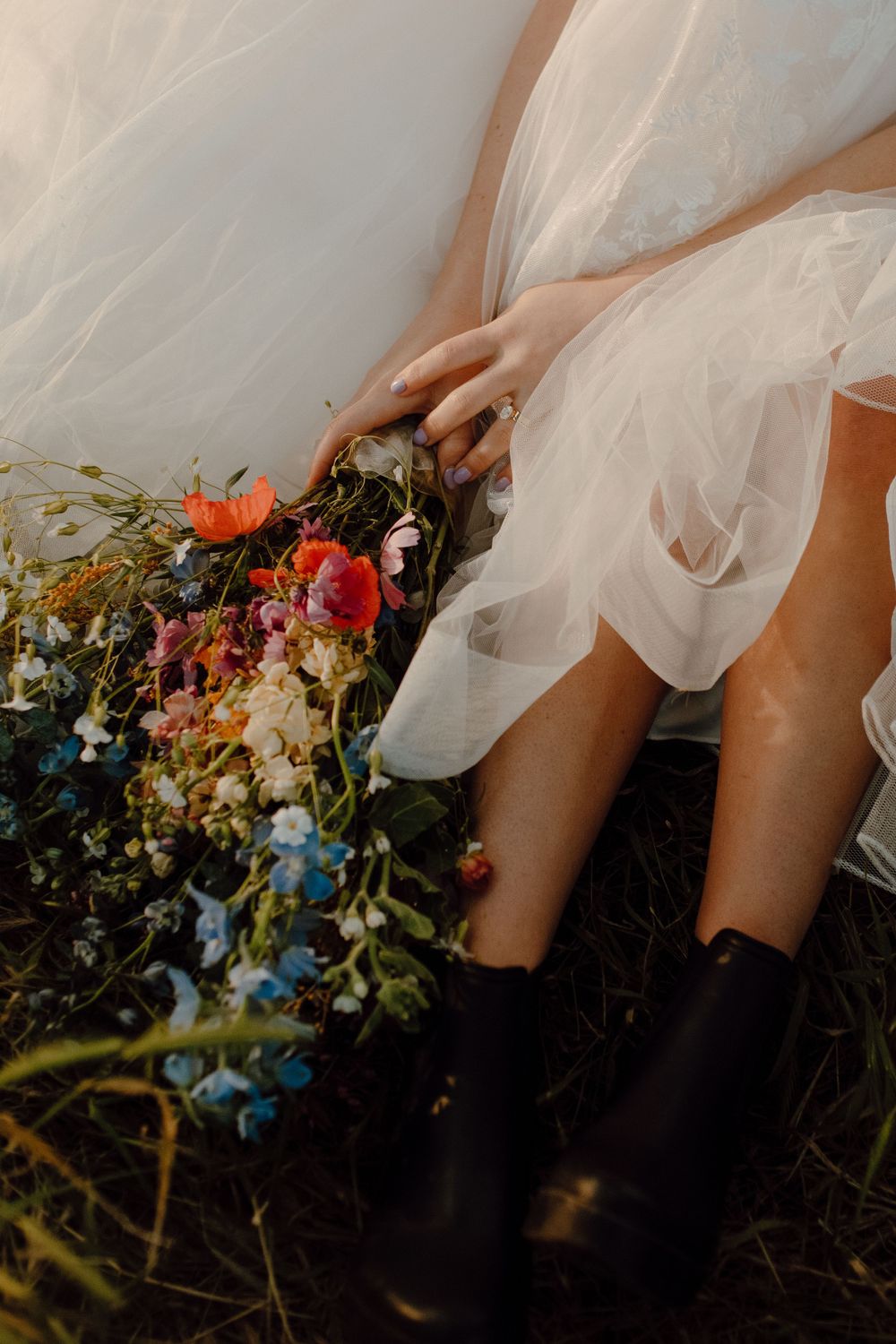 A feminine figure in a white tulle skirt and black boots sits with a colorful wildflower bouquet on the ground.