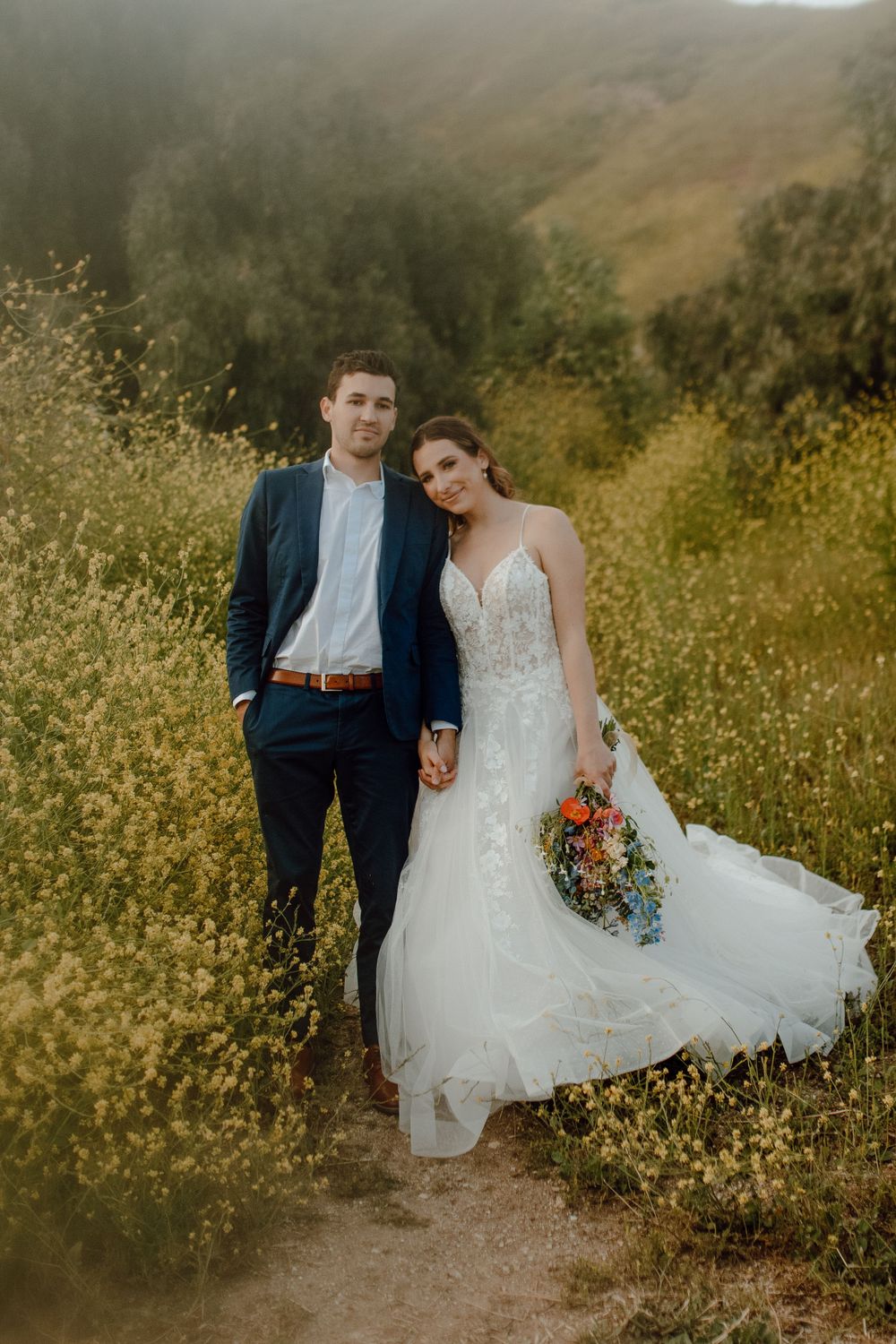 A couple in wedding attire poses together on a mountain trail surrounded by wildflowers during golden hour.