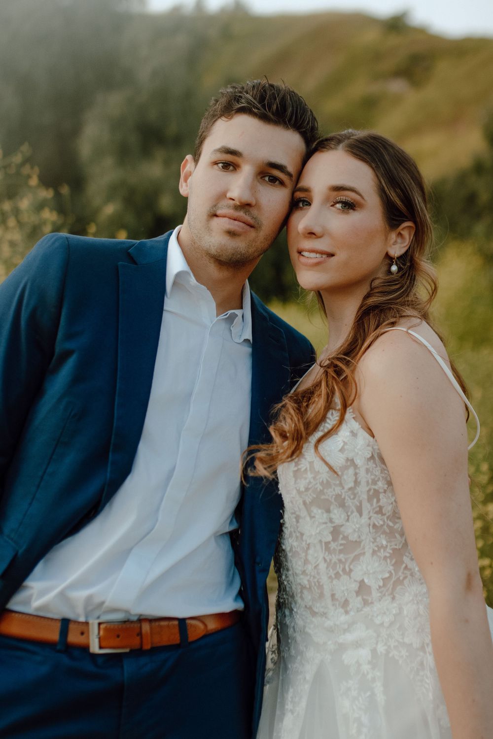 A romantic wedding portrait features a couple in a navy suit and delicate lace gown outdoors during golden hour.