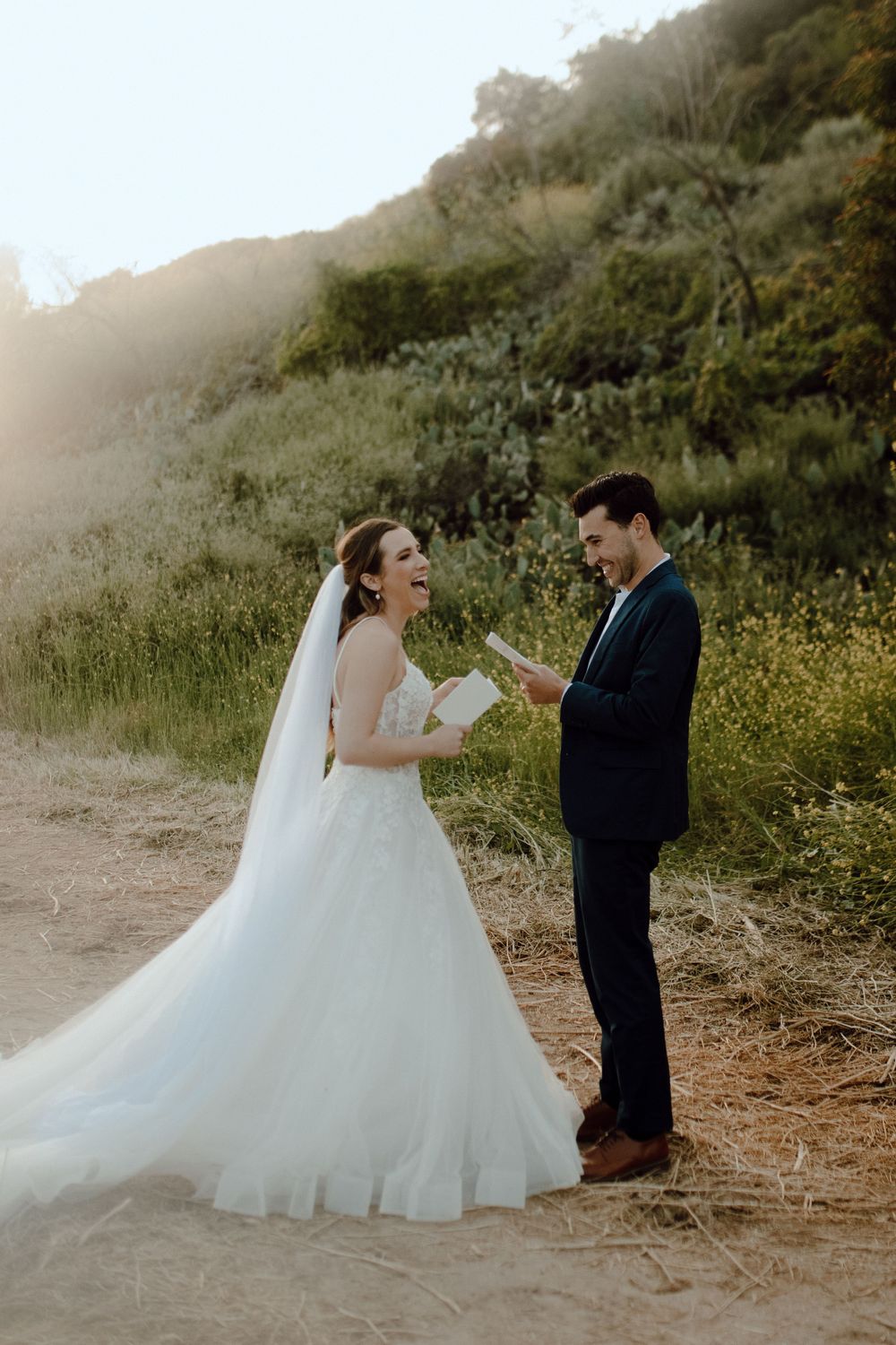 A couple in wedding attire shares an intimate moment on a sunlit dirt path surrounded by lush greenery.