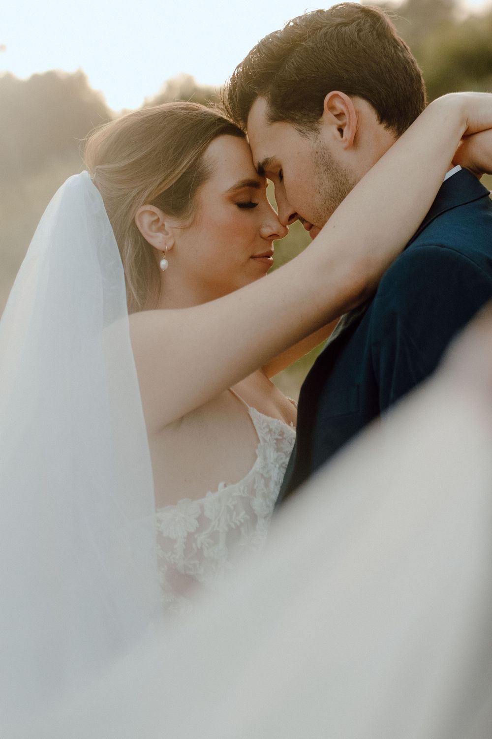A tender romantic moment between newlyweds in sunlight as a flowing white veil delicately frames their embrace.