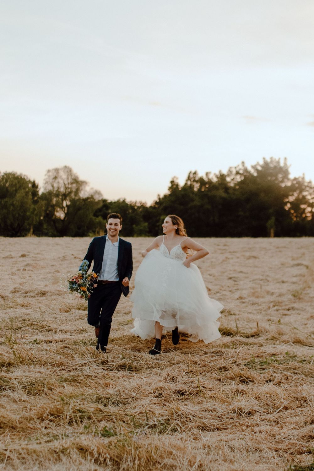 A couple in wedding attire runs playfully through a golden field at sunset while wearing sneakers.
