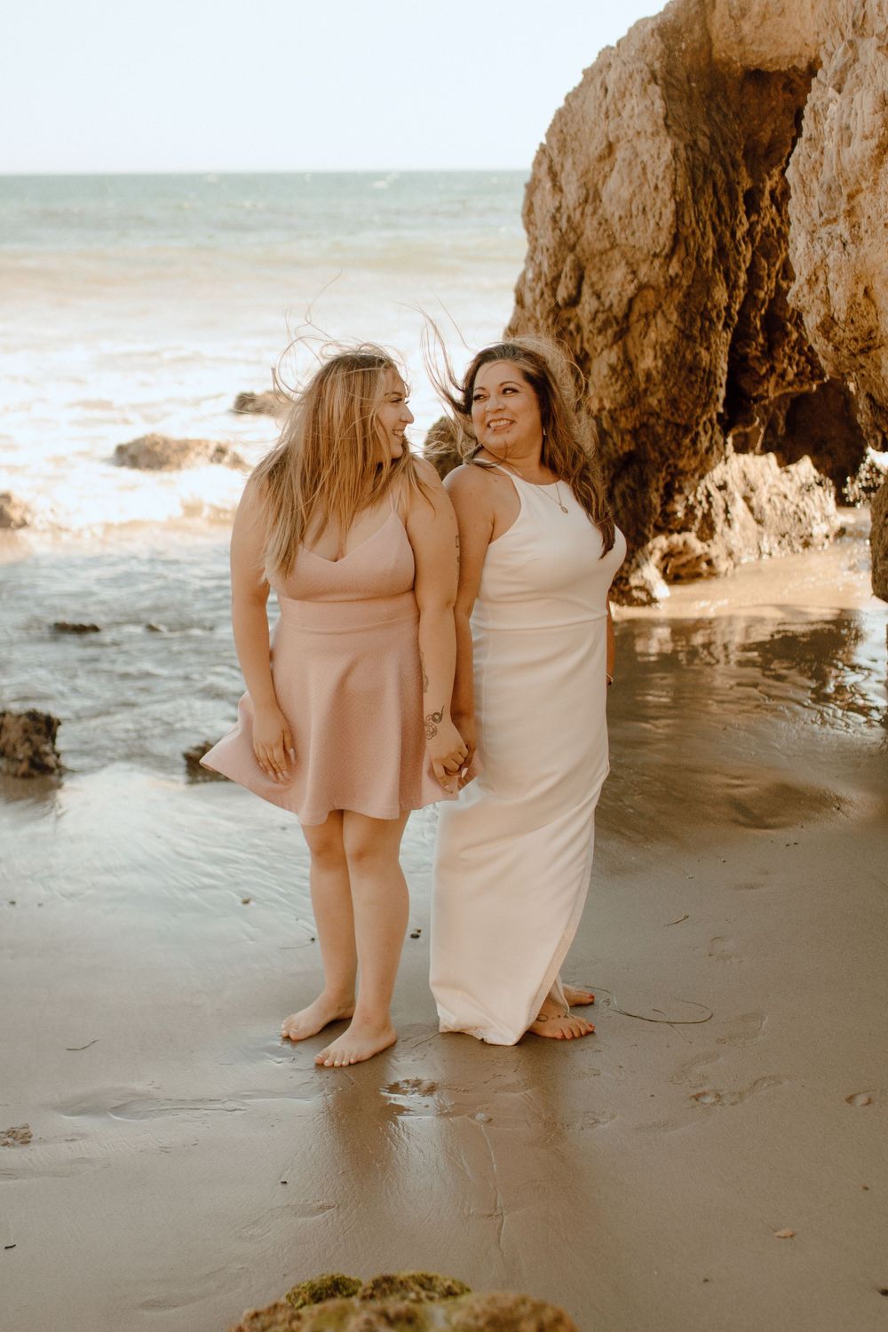 Two friends share a joyful moment together on a beach at sunset near rocky cliffs.