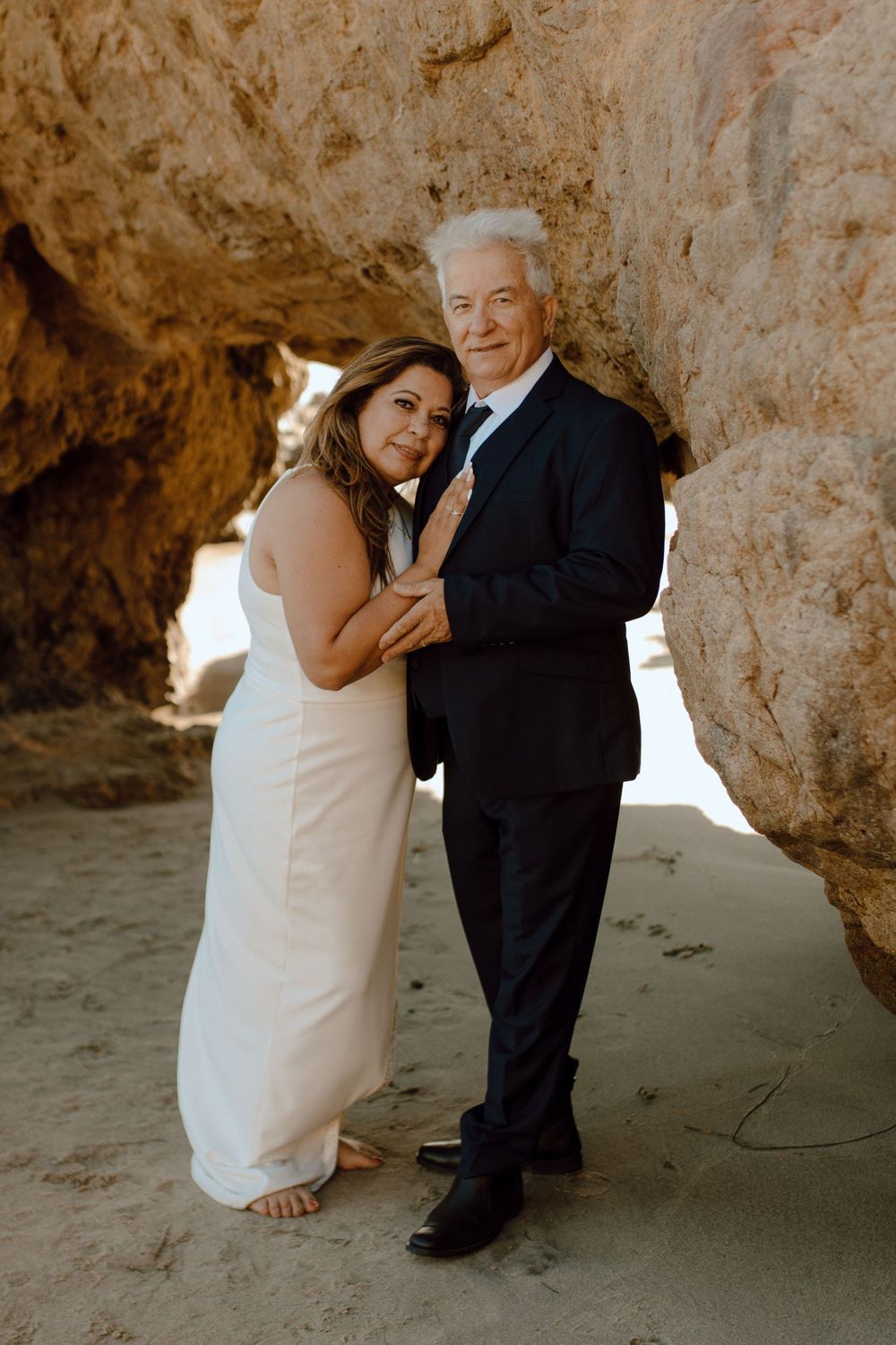 Couple embraces at a beach cave while wearing wedding attire during a romantic photoshoot.