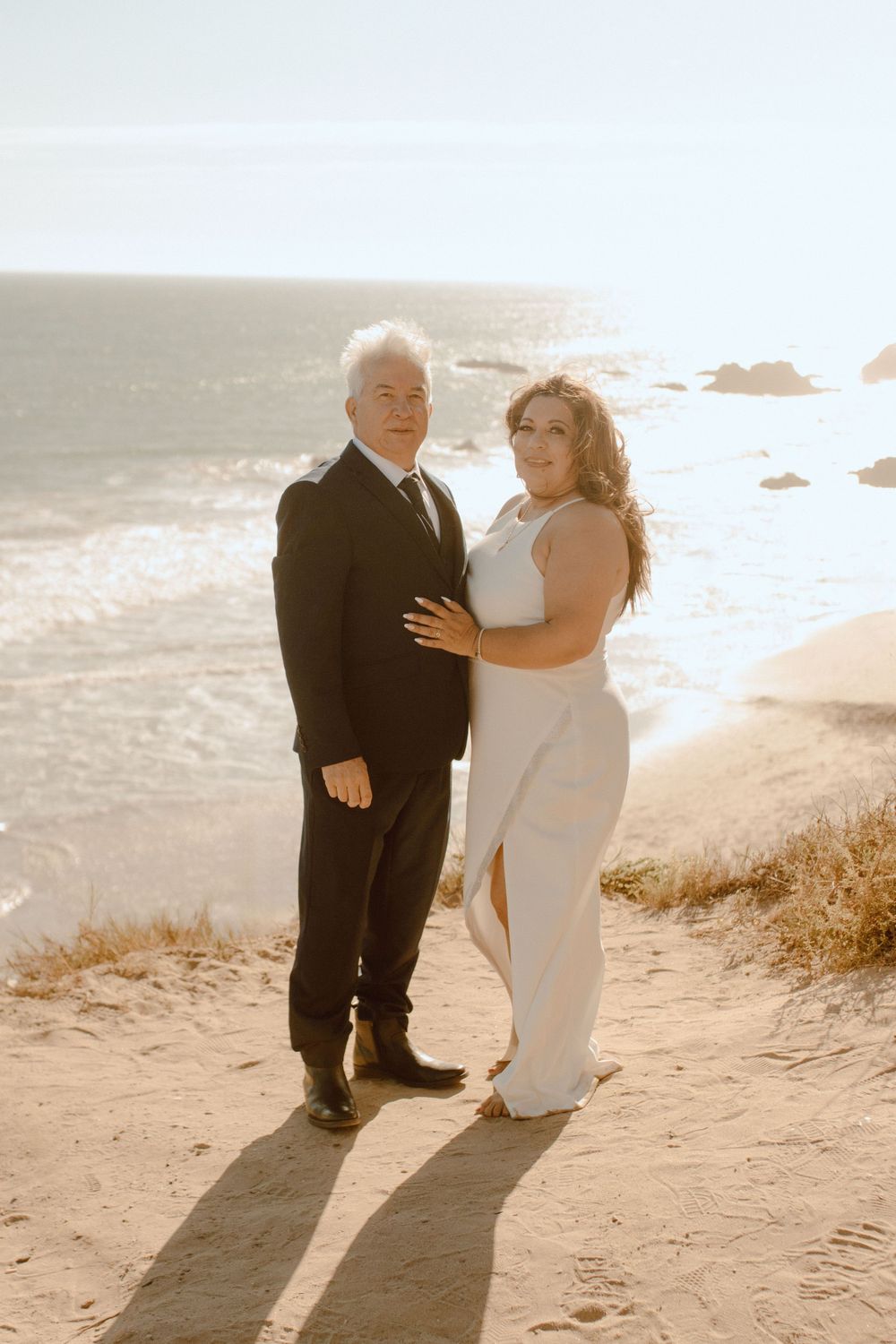 A couple in formal wedding attire embrace on a coastal cliff with dramatic ocean views at sunset.