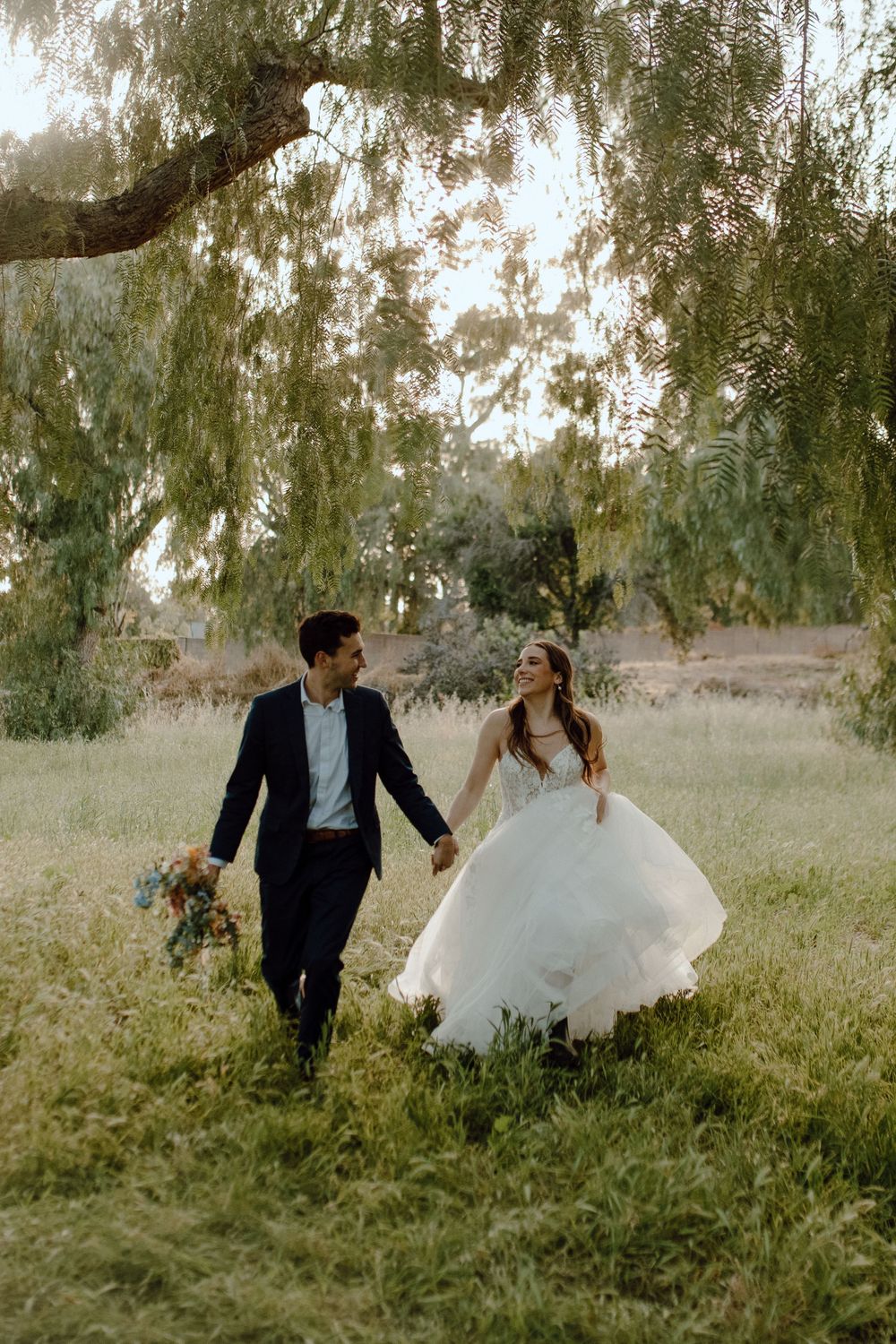 A couple in wedding attire holds hands while walking through a meadow beneath a willow tree on their special day.
