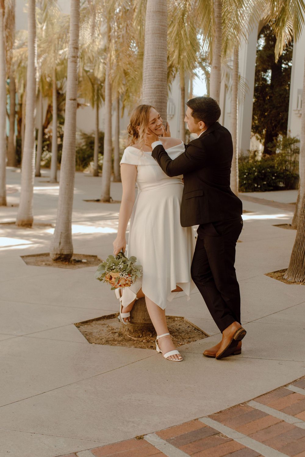 Romantic wedding couple shares an intimate moment while standing among palm trees on a brick sidewalk.