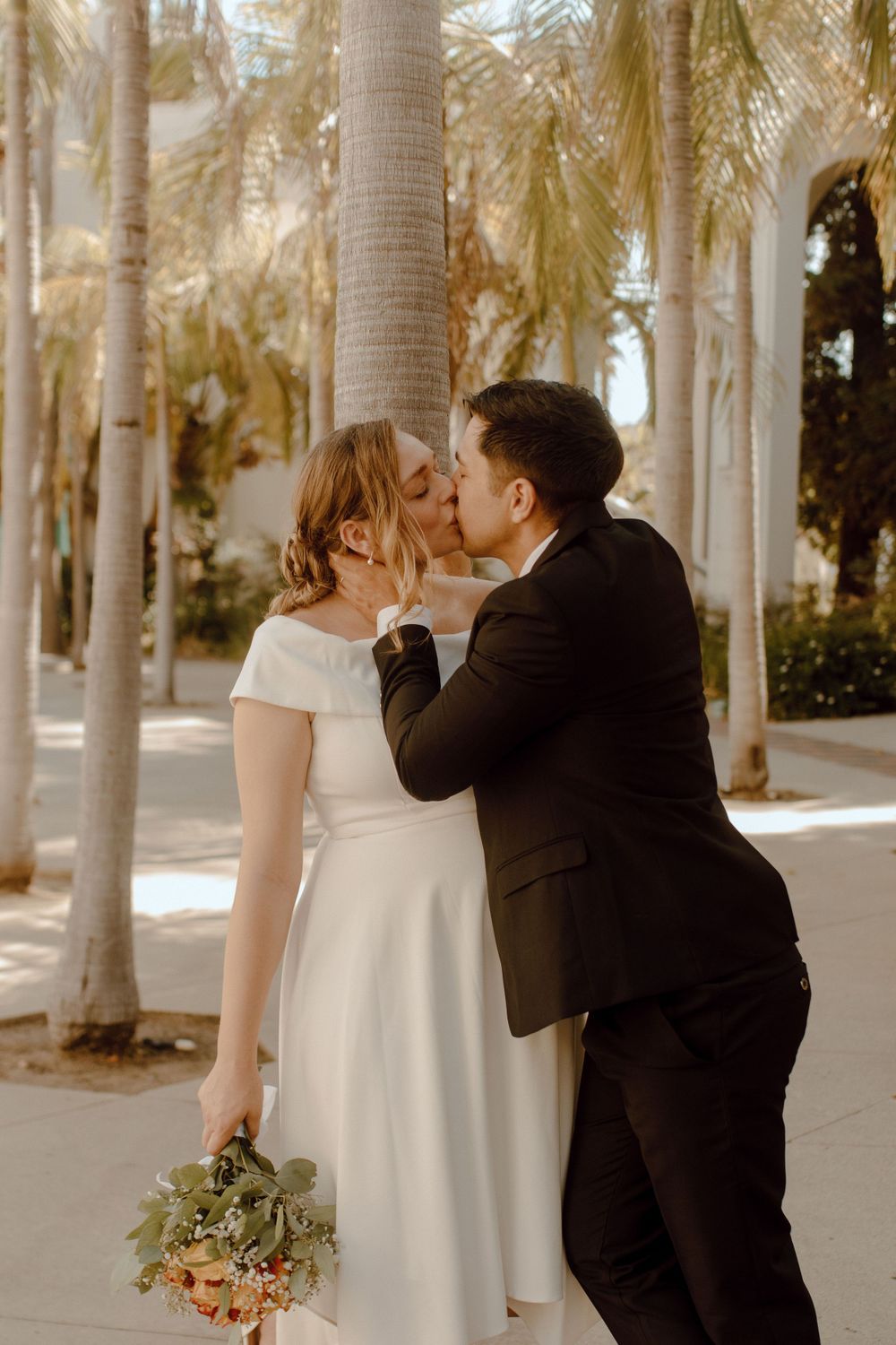 Couple shares a romantic kiss among palm trees during a golden hour wedding photoshoot.