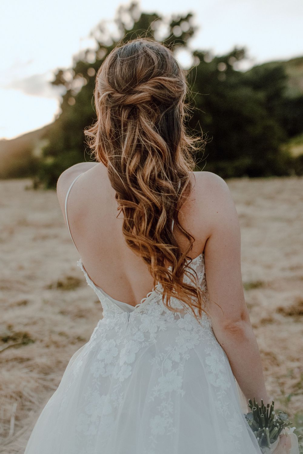 Back view of a bride in a white lace wedding dress with long wavy brown hair styled in romantic curls at sunset.
