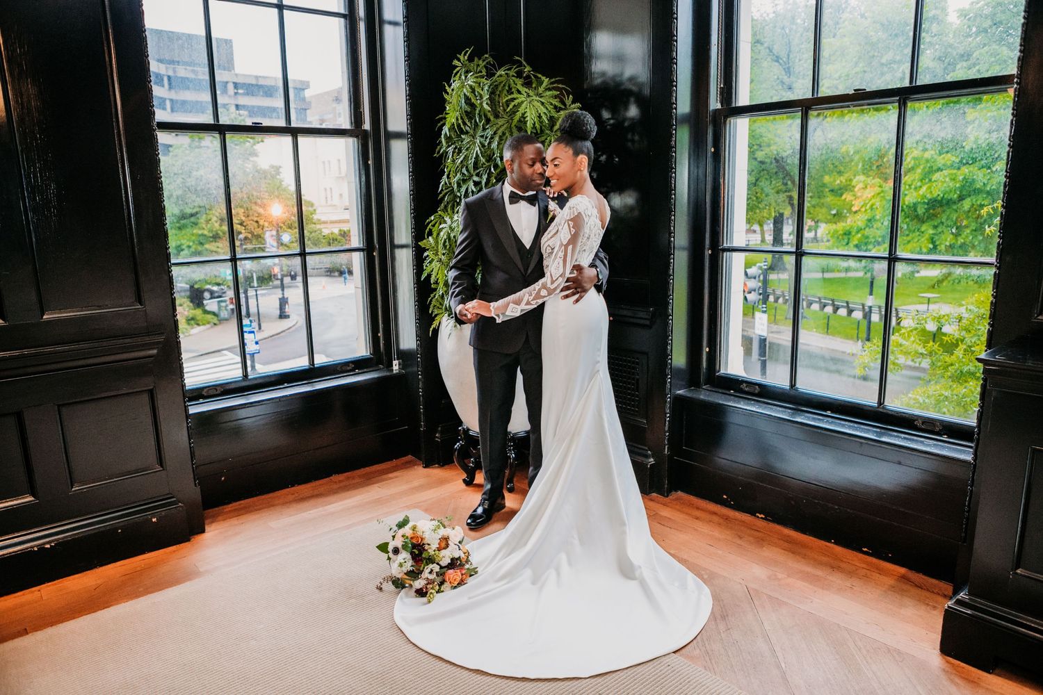 A romantic wedding moment captured in a sunlit corner between tall windows with garden views and a trailing white gown.