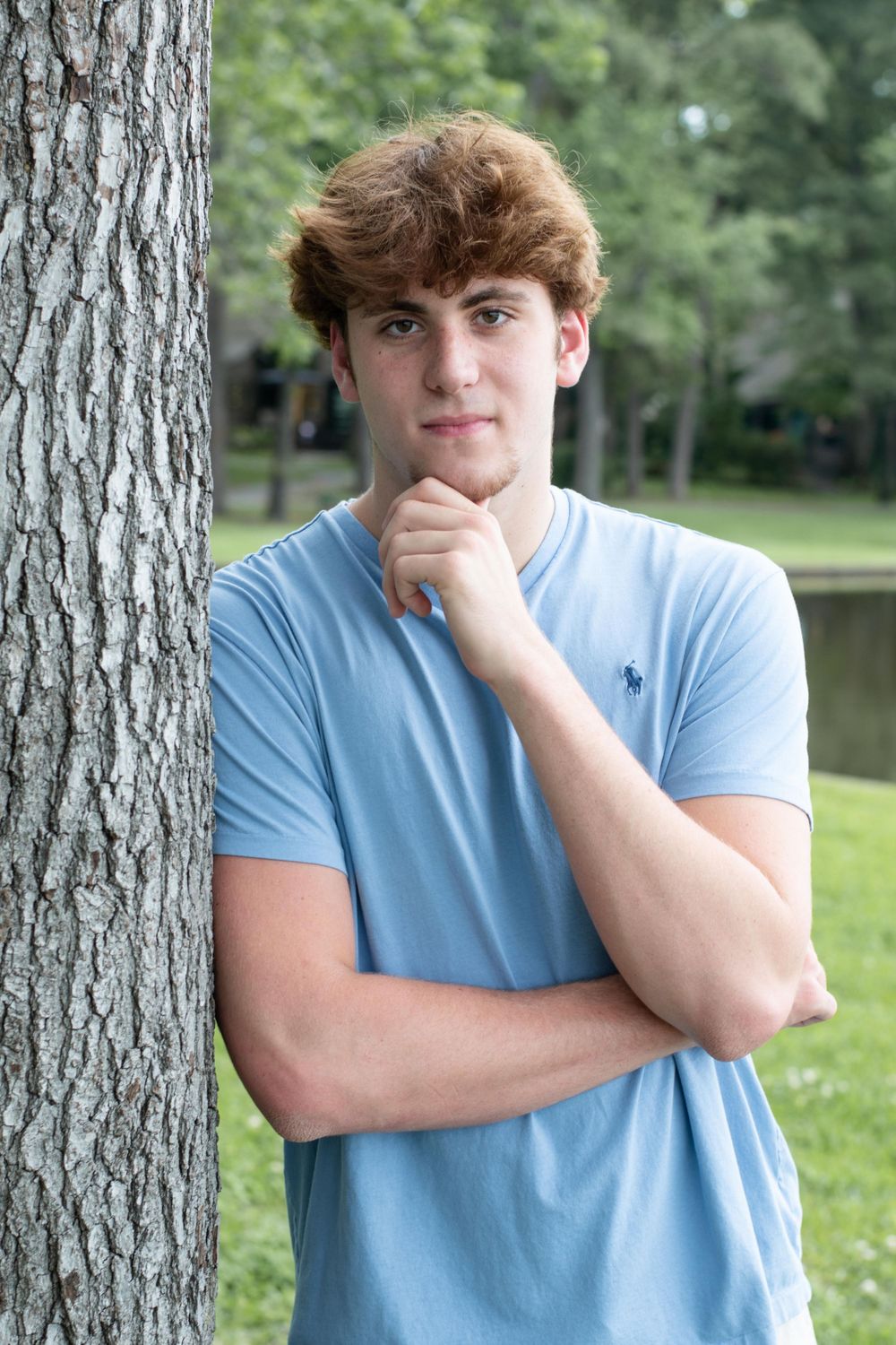Thoughtful pose against a tree trunk wearing a light blue t-shirt with a lake in the background.