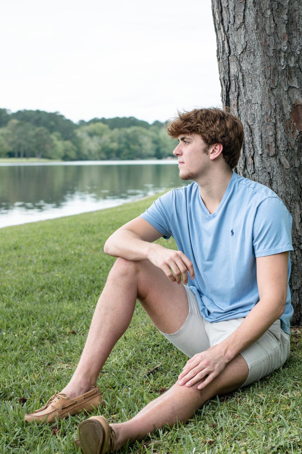 Person in summer attire relaxing on grass by a lake, leaning against a large tree.