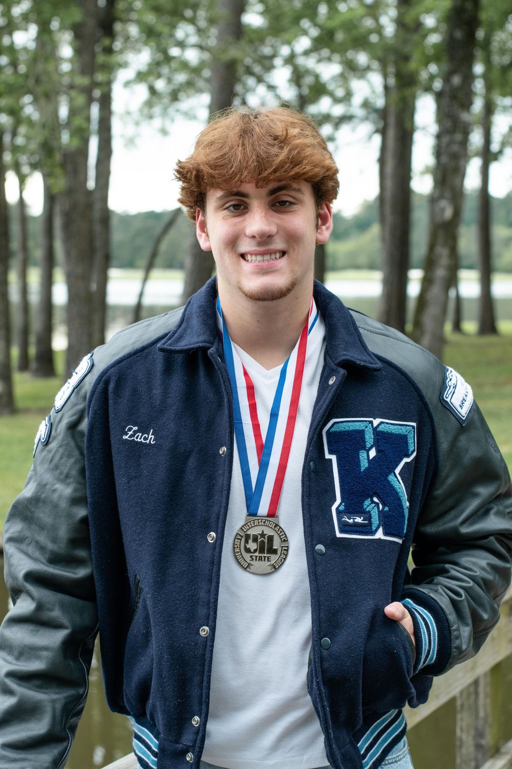 Proud athlete displays medal while wearing varsity jacket outdoors by scenic lake.