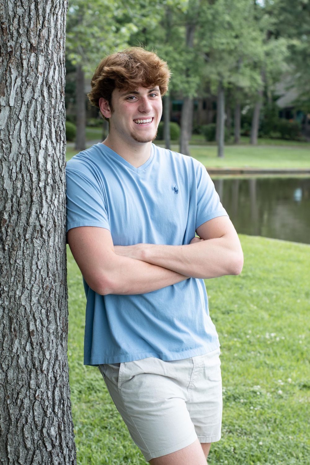 Athletic person in a light blue t-shirt poses confidently against a tree trunk near a serene lake.