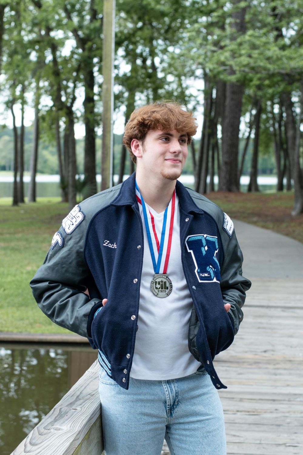 Student athlete wearing varsity jacket and medal poses on wooden boardwalk near lake.