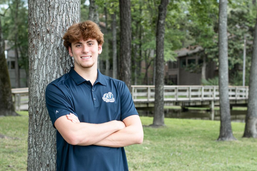 Athletic figure in a navy polo shirt poses confidently against a tree in a park setting.