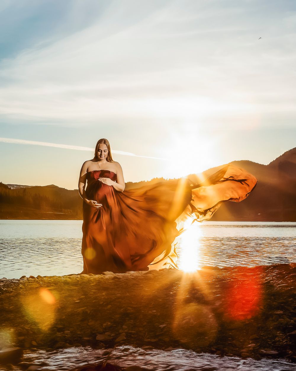 Gravid kvinne ved vannet med fjell og solnedgang i bakgrunnen, hun har på seg silkestoff som flyter i vinden