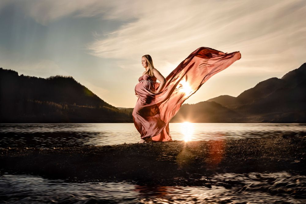 Gravid kvinne ved vannet med fjell og solnedgang i bakgrunnen, hun har på seg silkestoff som flyter i vinden