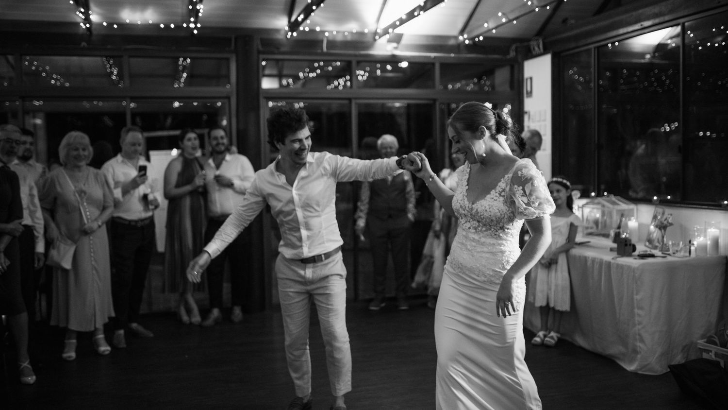 A couple shares their first dance at a wedding reception while guests look on in black and white.