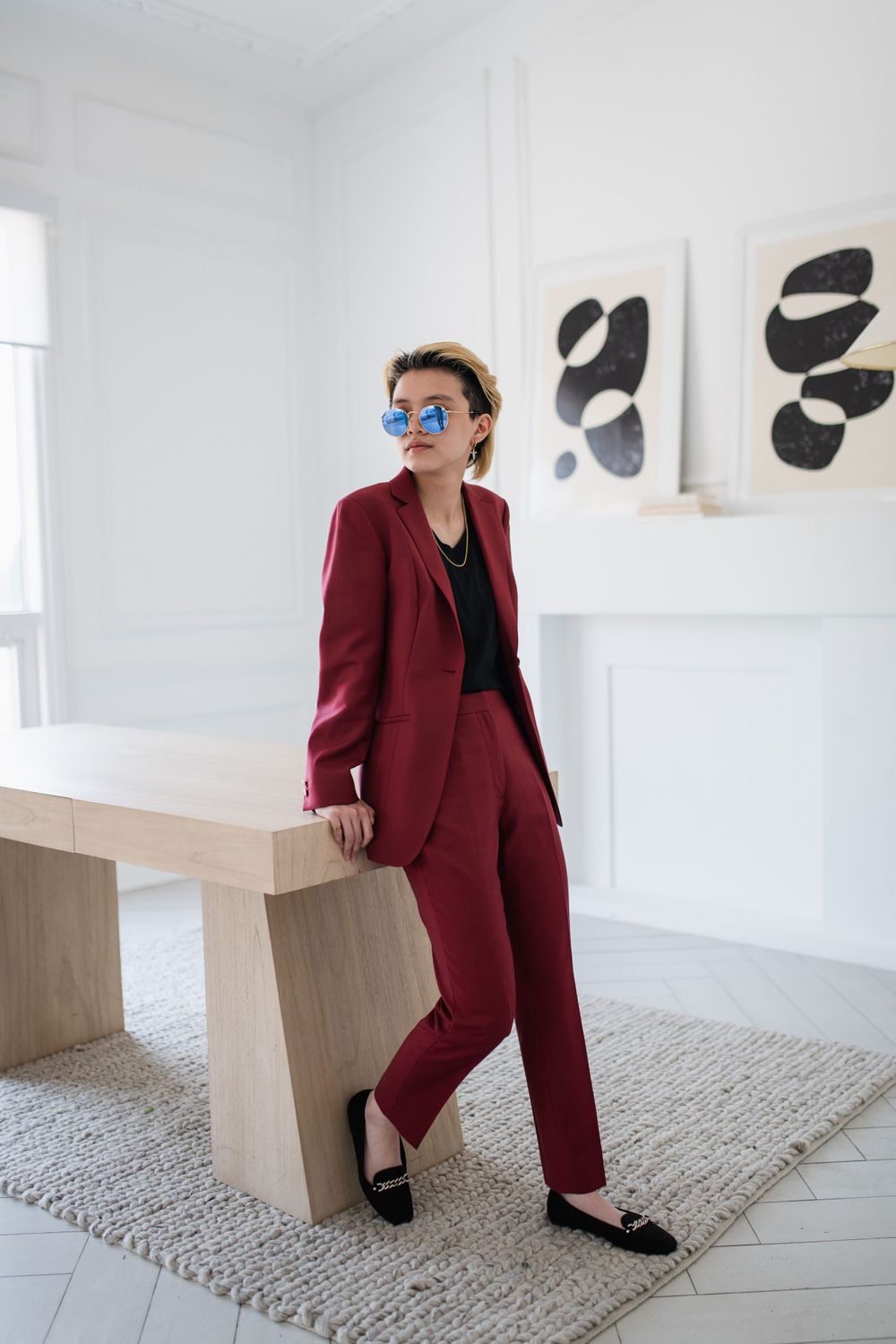 Stylish person in burgundy suit and sunglasses leans against wooden bench in modern white space.