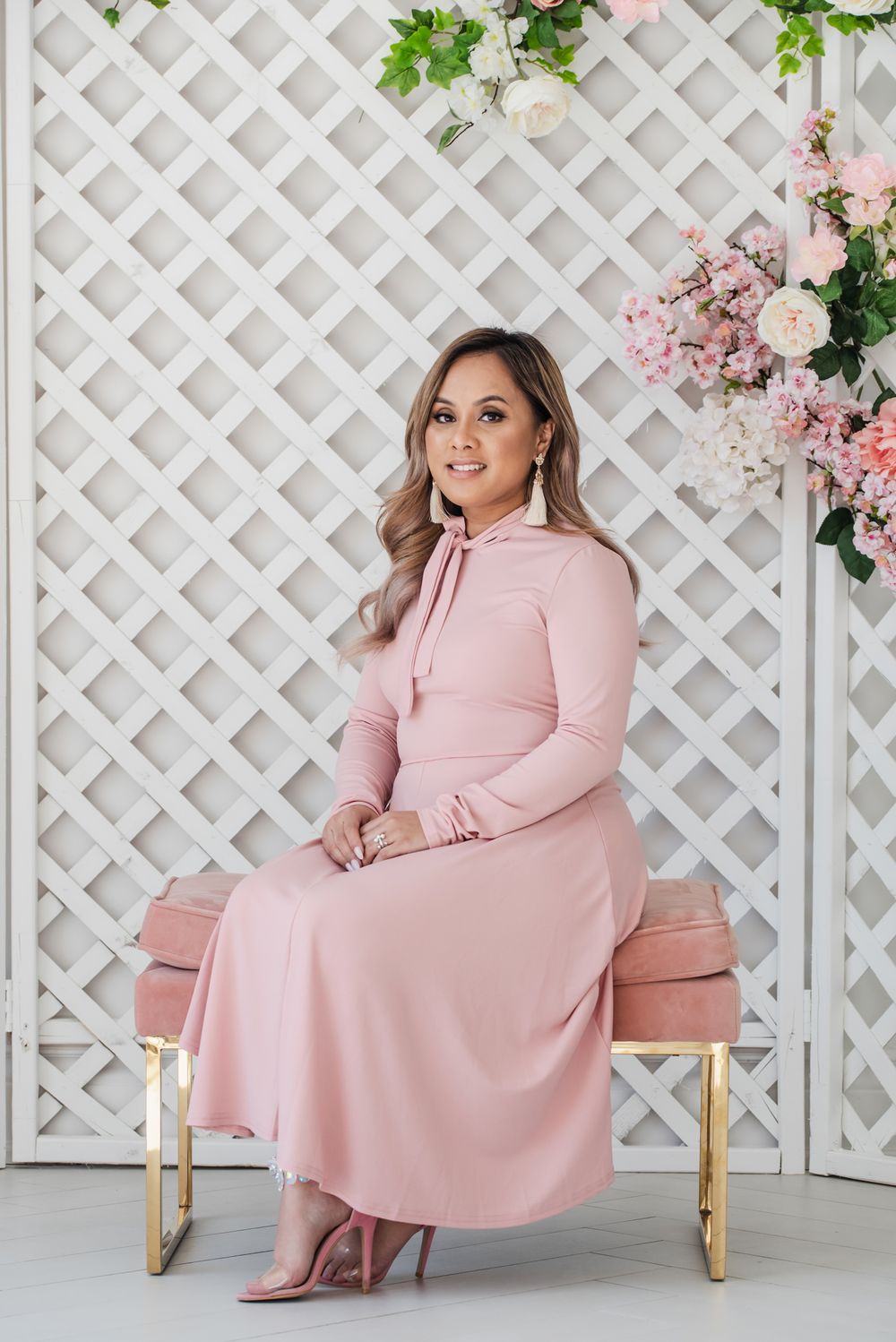 Woman in pink long-sleeve dress sitting on gold bench against white lattice backdrop with floral decorations.