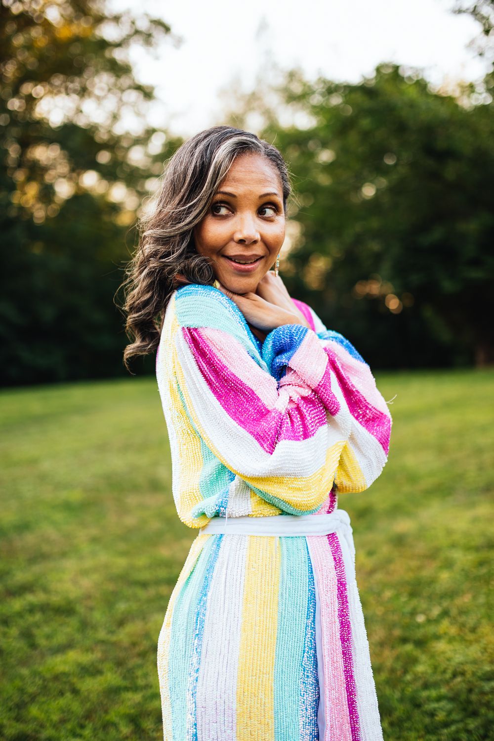 A person in a rainbow striped dress poses outdoors in nature with a joyful expression.
