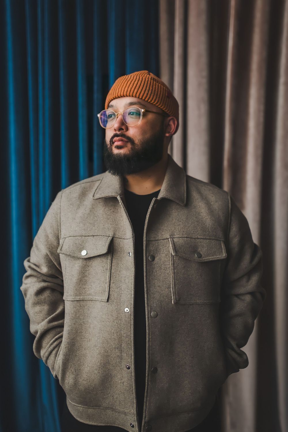 Man wearing orange beanie and gray jacket poses against blue and gray curtain backdrop.