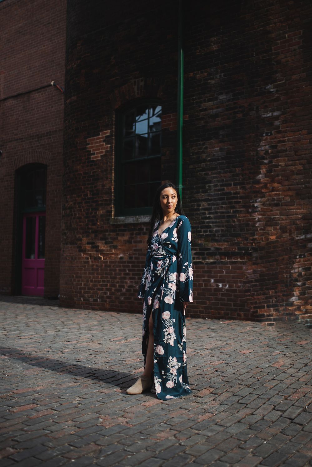 Woman in floral maxi dress poses against rustic brick wall on cobblestone street.