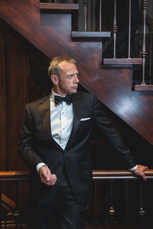 A person in a tuxedo poses dramatically against wooden stairs in moody lighting.