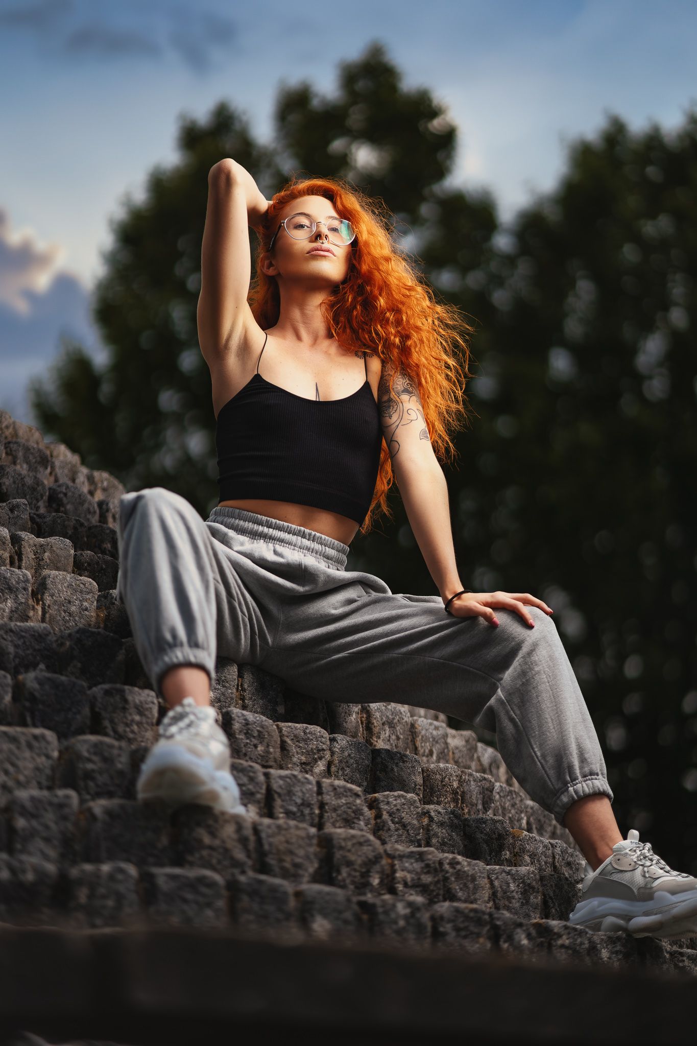 Red-haired woman seated on stone steps in a black crop top, full body fashion portrait at dusk with natural backdrop