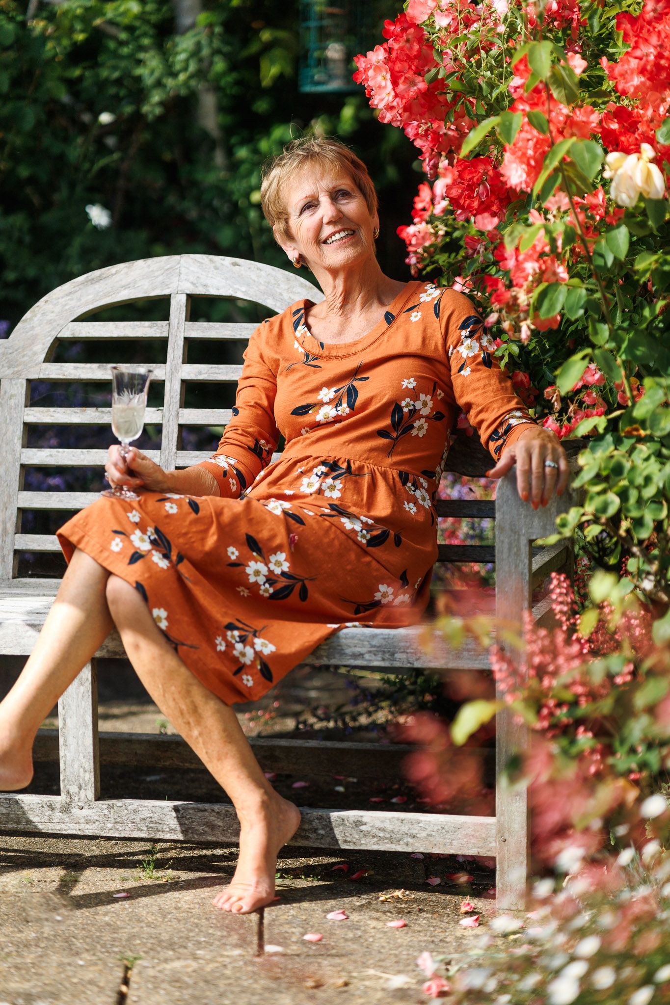 Woman in an orange floral dress on a garden bench with climbing roses, outdoor environmental portrait photography