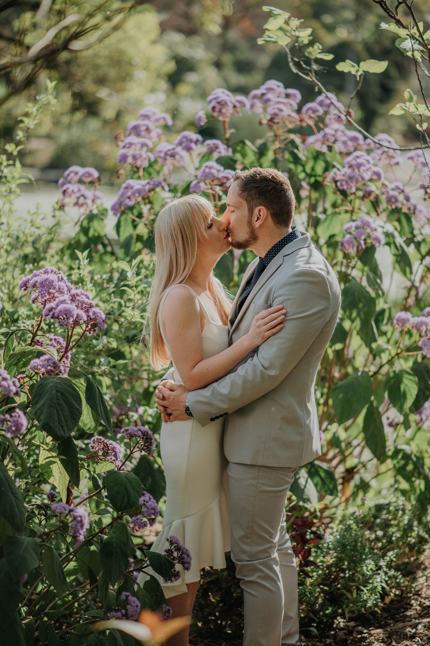 Couple shares intimate kiss among blooming purple flowers in garden setting.