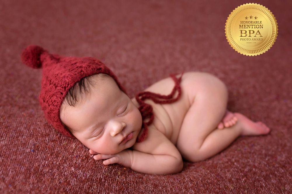 Sleeping newborn wearing red knit hat posed on burgundy backdrop.