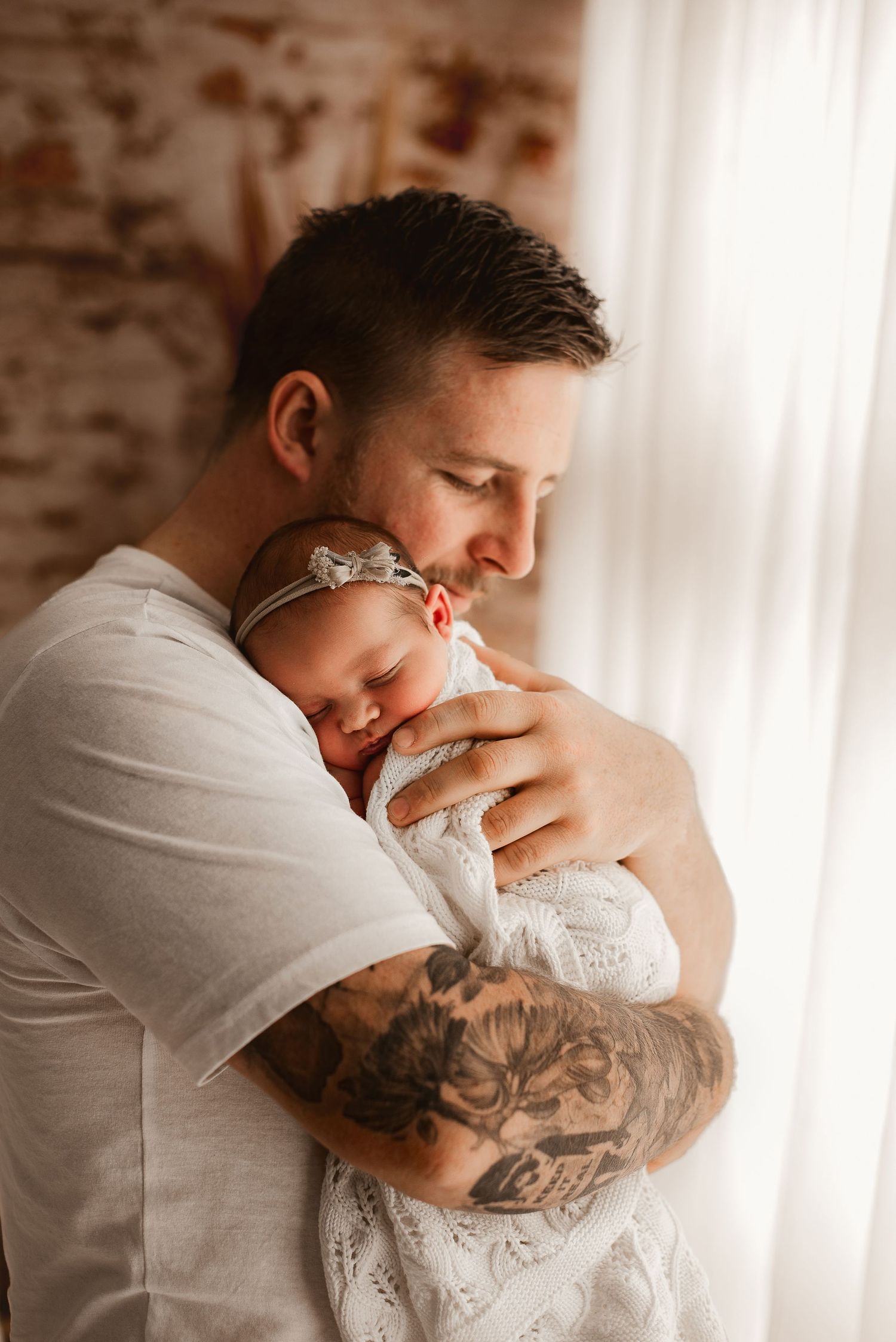 Parent with tattoo sleeve holding newborn baby near window with exposed brick wall.