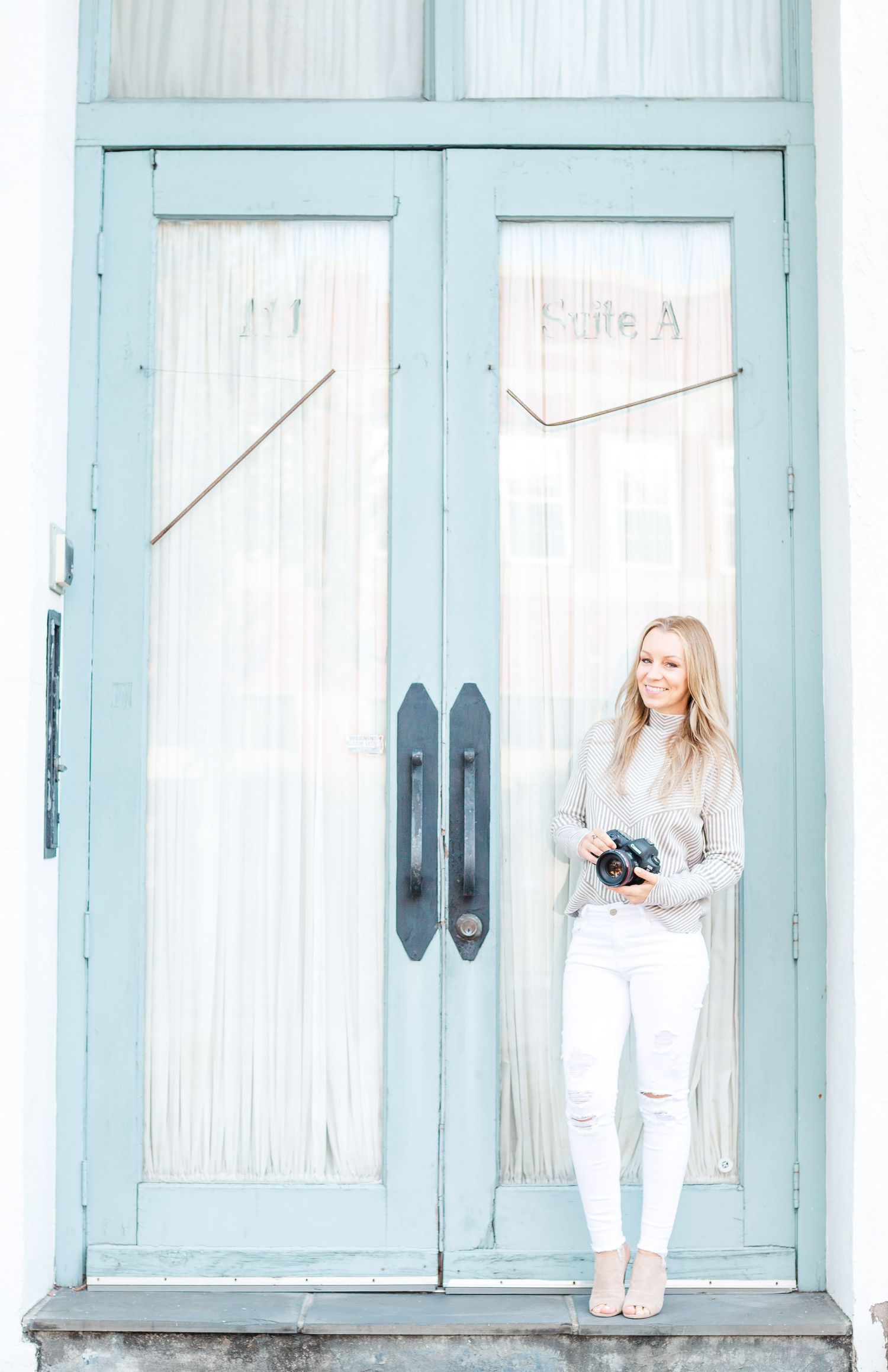 A figure in white stands against large vintage blue doors with white curtains.