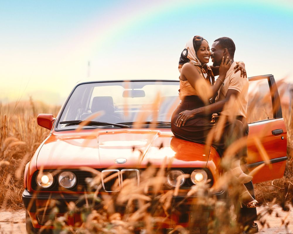 A romantic couple embraces in a vintage red convertible car with a rainbow in the background.