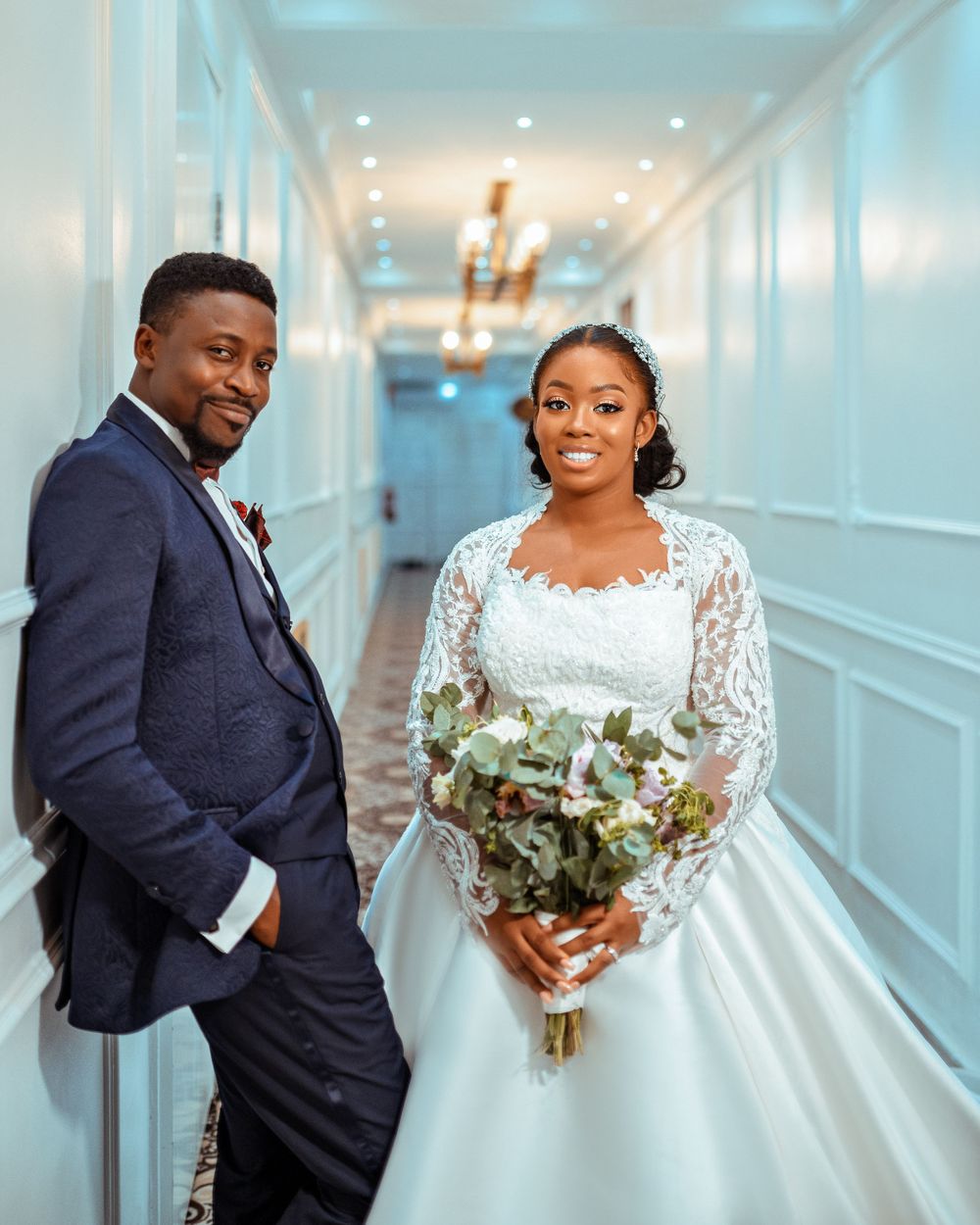 A wedding couple poses in an elegant hallway with the bride holding a bouquet of white flowers.