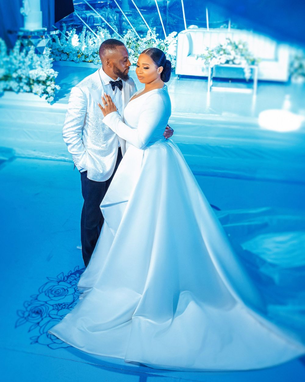 Wedding couple in white attire posed against blue-lit scenery.