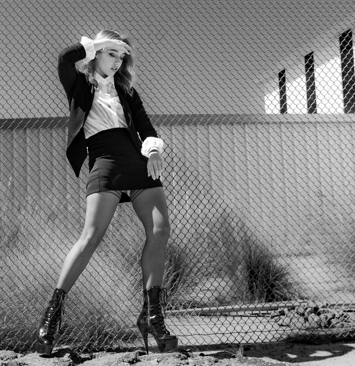 Black & white image of a stylist blonde woman wearing high heels and a black miniskirt, striking a provocative pose in front of a fence.