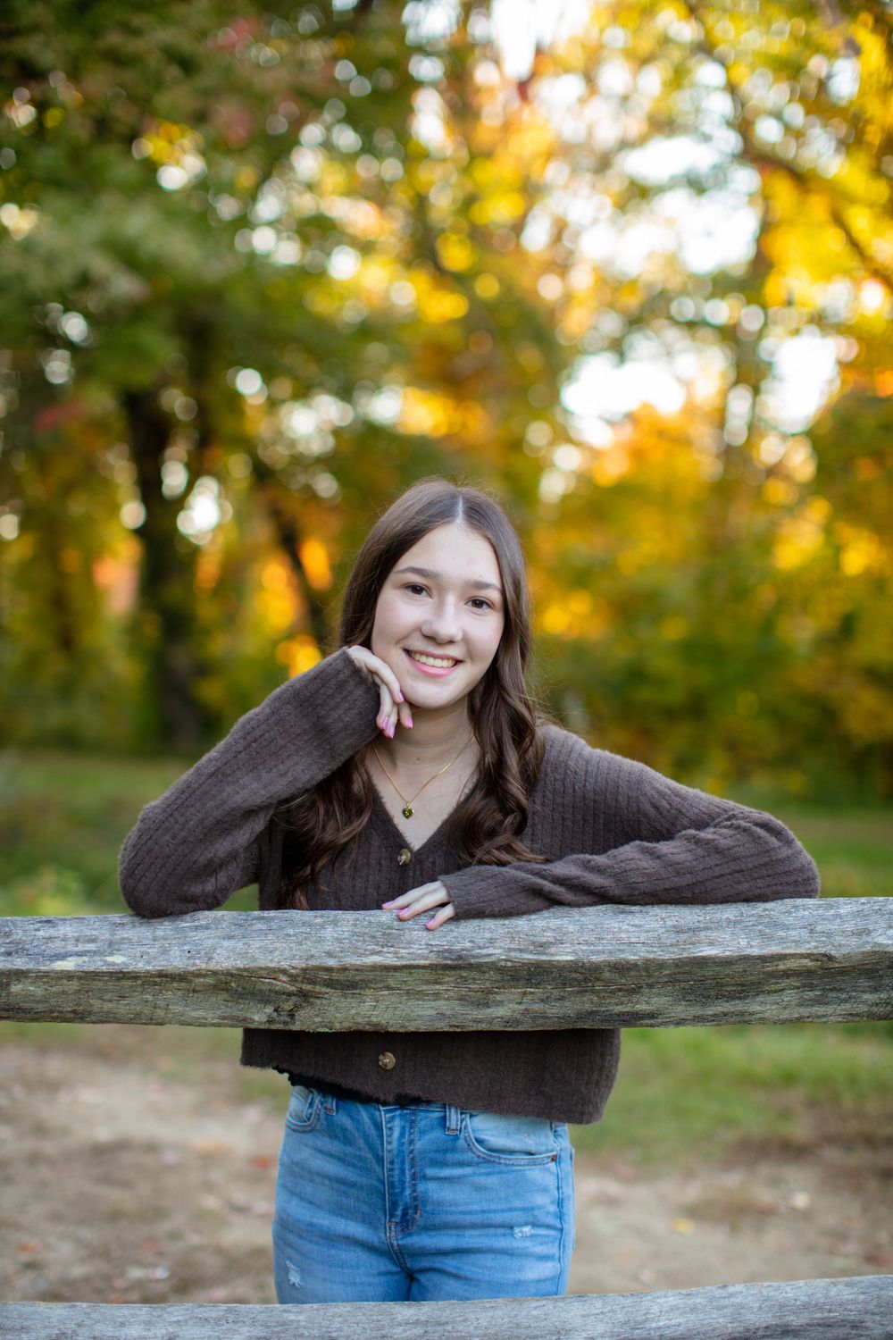 Person in brown sweater leaning on wooden fence with autumn foliage background.