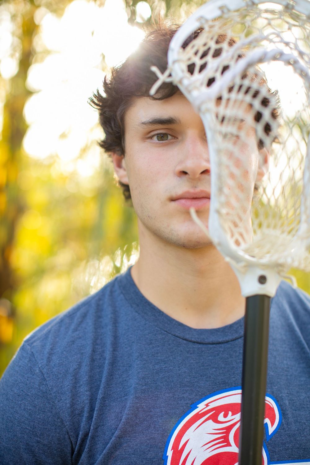 Athletic person in blue t-shirt holding lacrosse stick against autumn background.