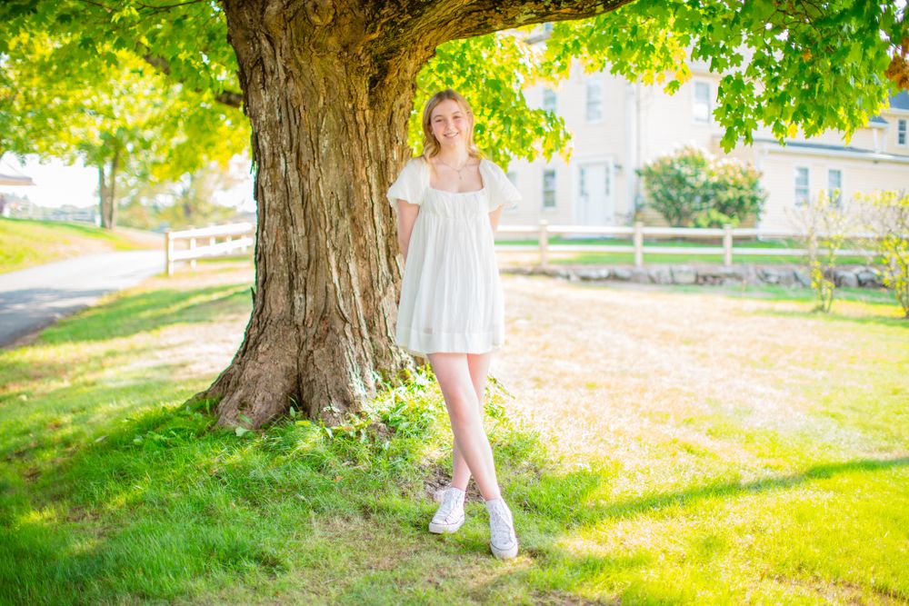 Person in white dress standing under large tree in sunny summer setting.