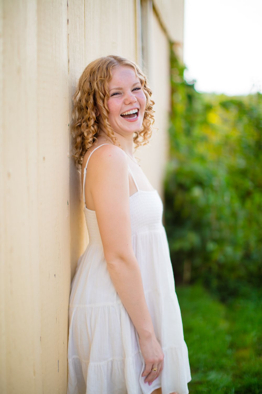 Person in white dress laughing joyfully while leaning against cream colored wall.