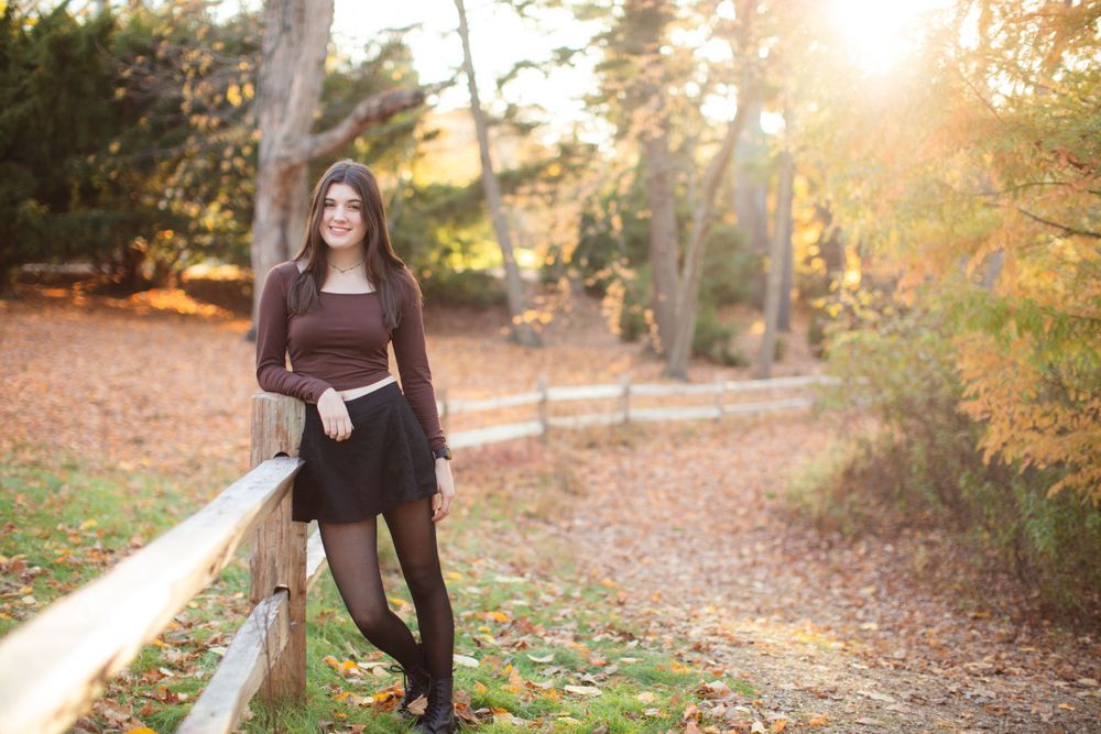 Person in brown dress leaning on fence during sunset in wooded park setting.