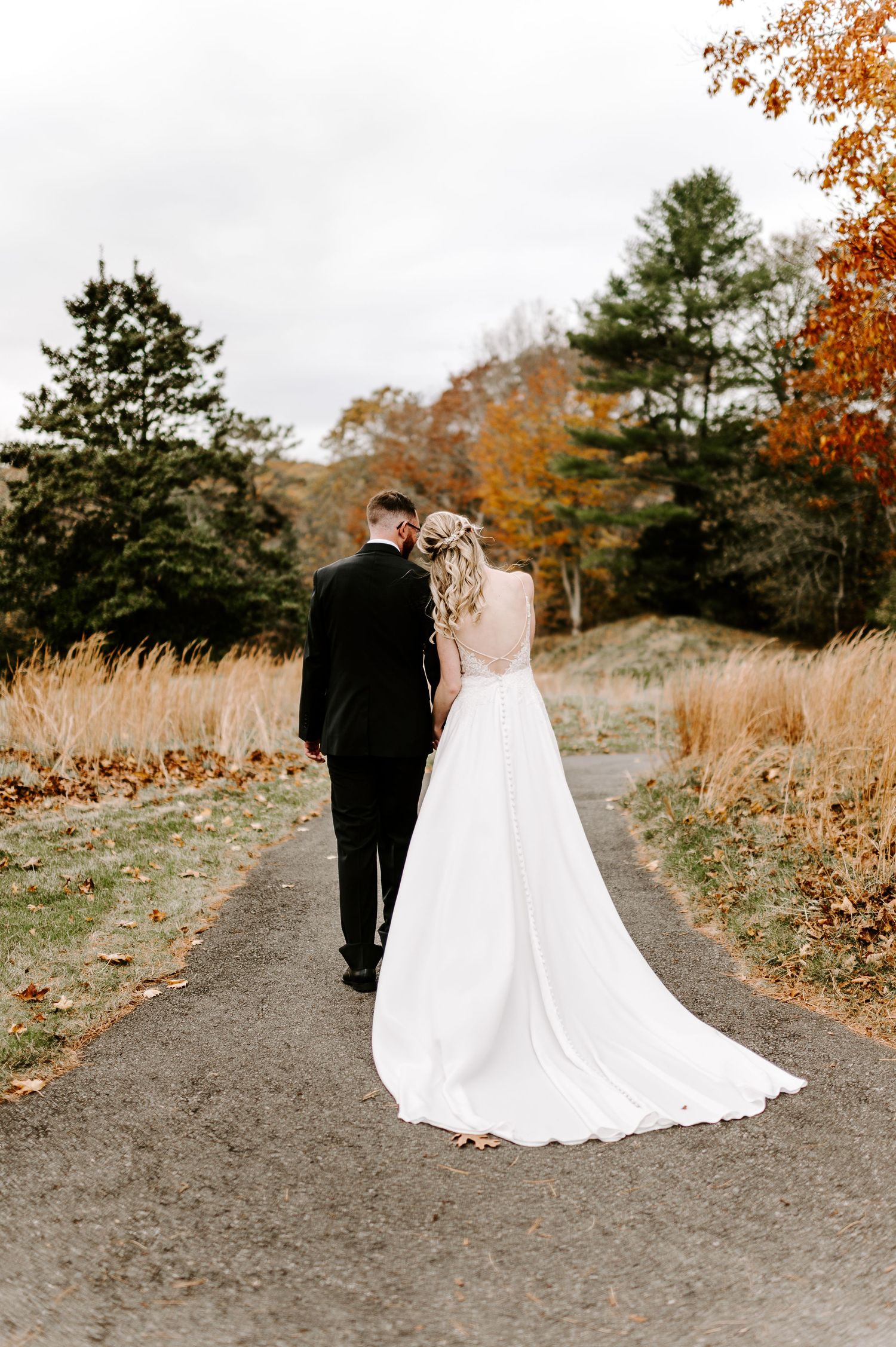 Bride in flowing white gown and groom walking down autumn path with golden grass.