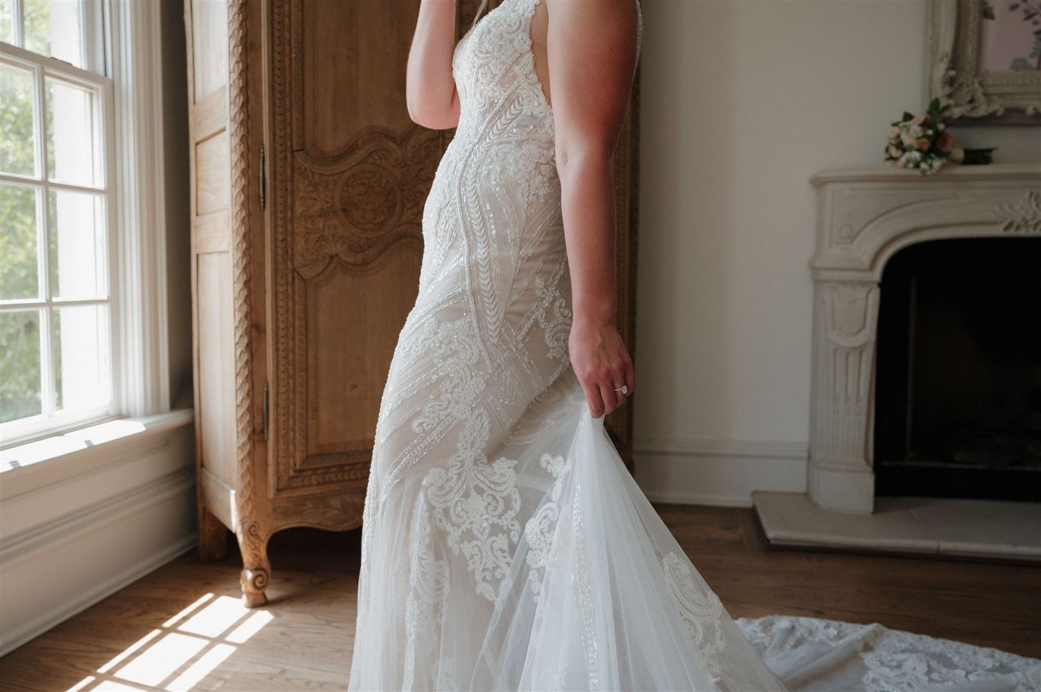 Sunlit detail of wedding dress train by window and vintage wooden armoire.